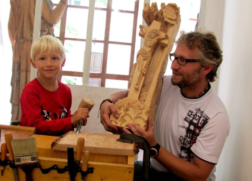 Šestiletý Ondřej Lukáš Vráblík vyřezává drobnou plastiku, vedle něj jeho otec Petr Vráblík třímá dřevěnou sochu, kterou vystavuje na trienále.