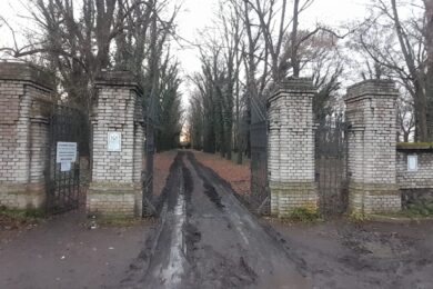 Vstupní brána do bohnického hřbitova – těžké železné křídlo mezi cihlovými pilíři otevírá cestu do místa, kde se historie mísí s legendami. Blátivá cesta lemovaná stromy připomíná, že za zdí se skrývá prostor tichých vzpomínek, zapomenutých osudů i dobrovolnického úsilí, které mu vrací důstojnost. Atmosféra zimního dne podtrhuje vážnost okamžiku: vstupujeme do areálu, který není atrakcí, ale místem paměti.