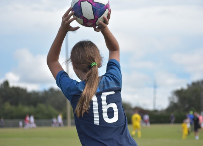 Mladá fotbalistka z Prahy 8 vhazuje míč zpět do hry. Symbol odhodlání, radosti z pohybu a práce klubů, které vedou dívky i chlapce ke sportu a zdravému životnímu stylu.