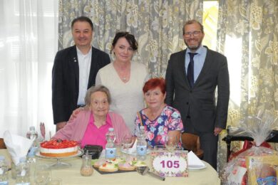 Slavnostní chvíle u narozeninového stolu. Paní Marie Lisá v růžovém svetru v kruhu gratulantů, s dortem a symbolickou číslicí 105.