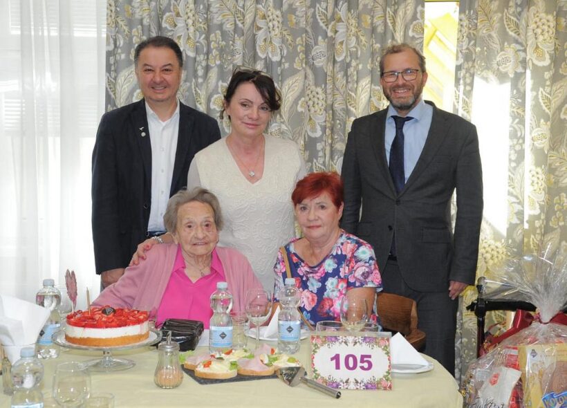 Slavnostní chvíle u narozeninového stolu. Paní Marie Lisá v růžovém svetru v kruhu gratulantů, s dortem a symbolickou číslicí 105.